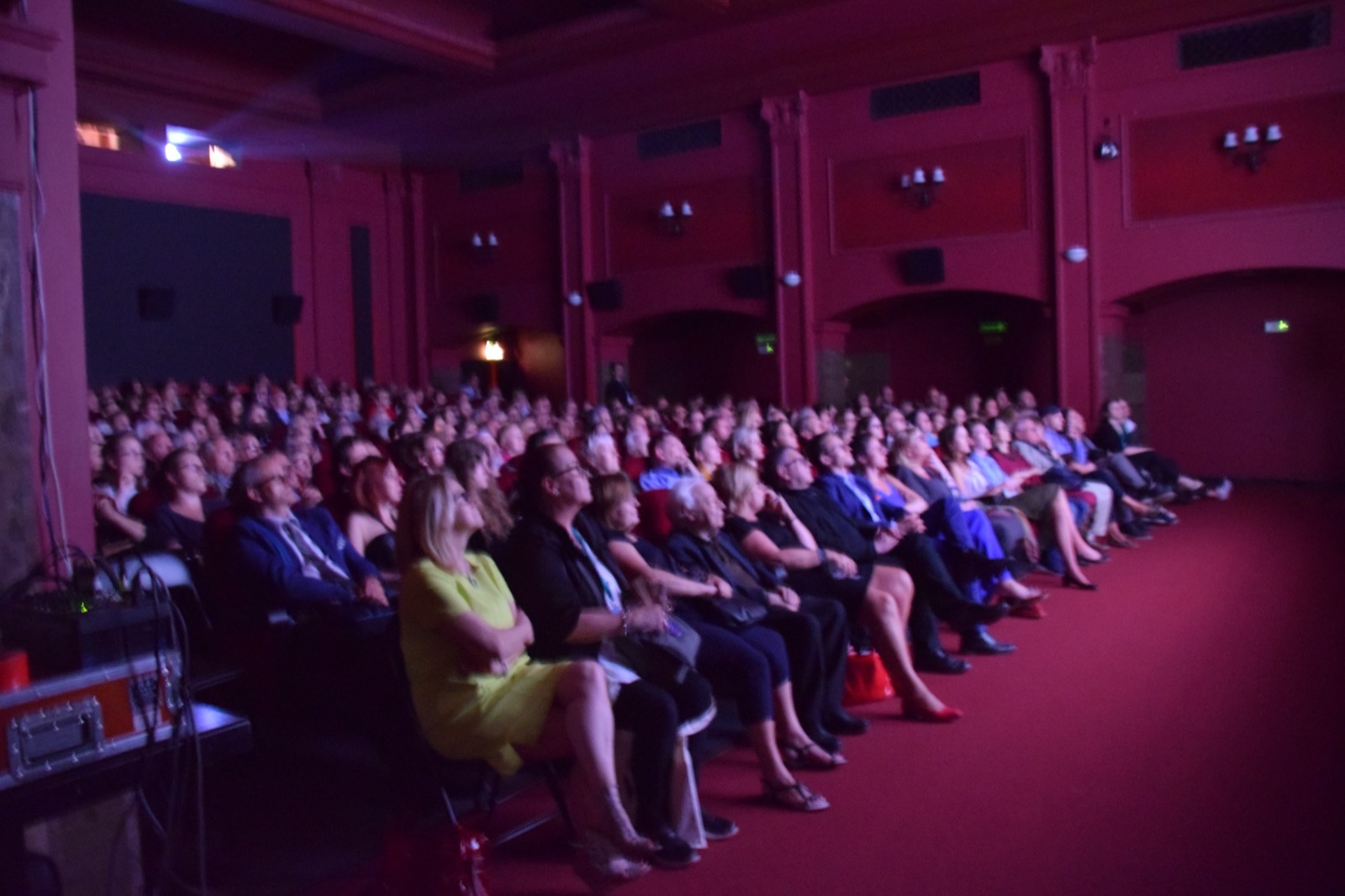 A full audience seated in a red-walled theater, watching the stage intently.